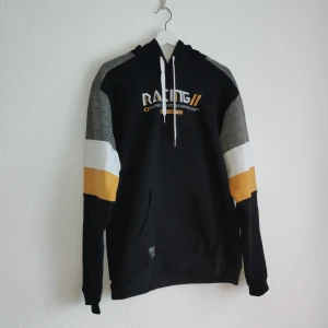 Svart hoodie från Pole Position - Säljer en svart hoodie från Pole Position med grå, vita och gula detaljer på ärmarna. Den har en justerbar huva med snören och texten 'Racing' tryckt på bröstet. Perfekt för en sportig look!
