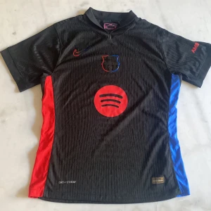 Svart FC Barcelona fotbollströja - Snygg svart FC Barcelona tröja från Nike med röda och blå detaljer. Tröjan har korta ärmar och klubbens emblem på bröstet. Perfekt för fans som vill visa sitt stöd med stil.