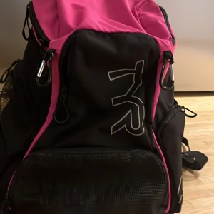 Svart och rosa ryggsäck från TYR - Säljer en snygg svart och rosa ryggsäck från TYR. Den har flera praktiska fack och dragkedjor för enkel förvaring. Perfekt för träning eller vardagsbruk. Ryggsäcken har justerbara axelremmar för bekväm passform.
