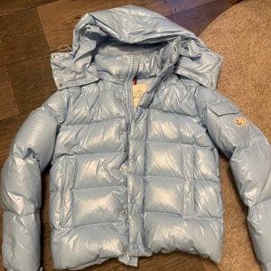 Moncler maya - Säljer en snygg ljusblå dunjacka från Moncler med en quiltad design och avtagbar huva. Jackan har en dragkedja framtill och tryckknappar. Den är dekorerad med Moncler-logotypen på ärmen och har en unik serietidningsetikett på insidan. St:2 motsvarar small/medium priset ej hugget i Sten kom med frågor och funderingar.