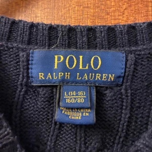 Mörkblå stickad tröja från Ralph Lauren - Säljer en snygg mörkblå stickad tröja från Ralph Lauren med det klassiska röda logotypbroderiet på bröstet. Tröjan har en kabelstickad design och långa ärmar, perfekt för kyligare dagar. Passar både till jeans och chinos för en stilren look.