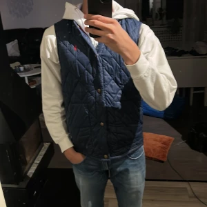 Blå quiltad väst från Polo Ralph Lauren - Säljer en snygg blå quiltad väst från Polo Ralph Lauren. Västen har en klassisk design med knappar och ett broderat rött märke på bröstet. Perfekt för lager-på-lager stil.