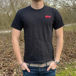 Svart t-shirt från Comme des Garçons - Säljer en svart t-shirt från Comme des Garçons med två röda hjärtan på bröstet. T-shirten är kortärmad och har en klassisk rund halsringning. Perfekt för en stilren look. Modellen är 180 cm liten i storleken. Köpt seconhand så ingen äkthet garanteras!