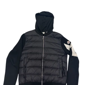 Svart pufferjacka från Moncler - Snygg svart pufferjacka från Moncler med huva och dragkedja. Jackan har långa ärmar med ett coolt vitt mönster och Moncler-logga på ärmen. Perfekt för kyligare dagar.