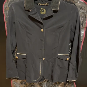 Svart tävlings kavaj från Tattini - Elegant svart kavaj från Tattini med gulddetaljer och kedja i ryggen. Kavajen har knappar framtill och på fickorna, samt kontrasterande piping längs krage och fickor. Perfekt för en stilren look. Medföljer väska 