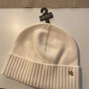 Vit stickad mössa från Lauren Ralph Lauren - Snygg vit stickad mössa från Lauren Ralph Lauren med ribbad kant och ett elegant guldemblem med RL-logga. Perfekt för att hålla värmen med stil.