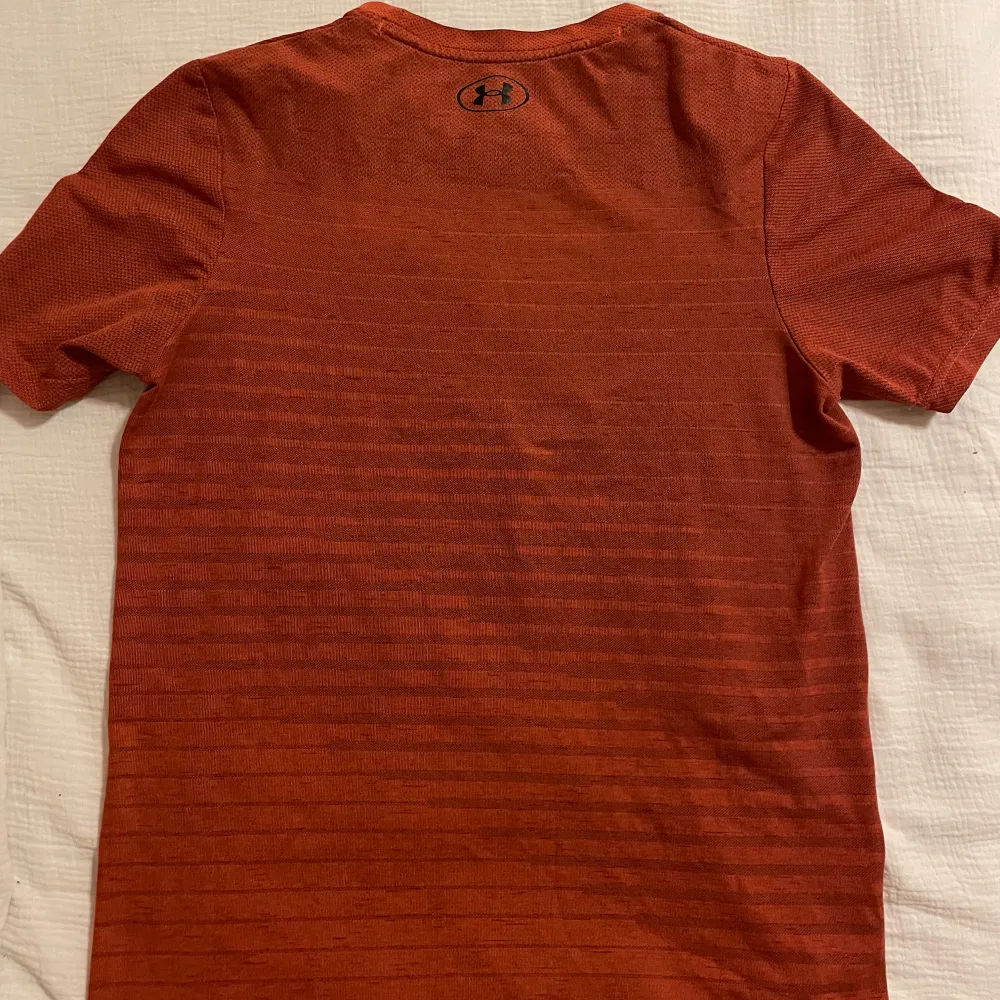 Säljer en snygg röd t-shirt från Under Armour med diskret randigt mönster. Perfekt för träning eller vardagsbruk. Den har korta ärmar och är tillverkad i ett lätt och bekvämt material. Passar en S/M.. Sportswear.