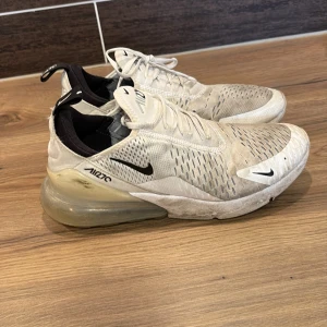 Nike Air Max 270  - Säljer ett par Nike Air Max 270 sneakers i beige med svarta detaljer. Skorna har en synlig luftkudde i hälen och snörning framtill. Perfekta för en sportig look.