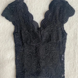 Svart spetsblus - Elegant svart spetsblus med v-ringad design och korta ärmar. Blusen har ett vackert mönster och en figurnära passform som framhäver silhuetten. Perfekt för en stilfull look.