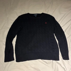 Mörkblå stickad tröja från Polo Ralph Lauren - Säljer en stilren mörkblå stickad tröja från Polo Ralph Lauren. Tröjan har ett diskret kabelstickat mönster och en liten röd logotyp på bröstet. Perfekt för kyligare dagar.
