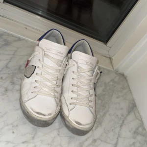 Vita sneakers från Philippe Model - Snygga vita sneakers från Philippe Model med blå och röda detaljer. Skorna har snörning och en unik design med gråa inslag på sidorna. Perfekta för en stilren look.