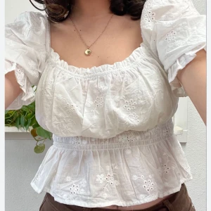 Vit blus med broderade detaljer - Charmig vit blus med broderade blommor och elastisk midja. Blusen har korta puffärmar med knytband och en söt volangkant nedtill. Perfekt för en somrig look! Mycket bra skick då den endast använts ett fåtal gånger. 