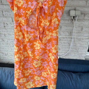 Orange blommig klänning med knyt - Säljer en färgglad orange klänning från H&M med blommigt mönster i gult och lila. Klänningen har smala axelband och en knytning framtill som ger en söt detalj. Perfekt för sommarens varma dagar!🌸💕🌸