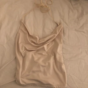 Fest topp - Säljer ett stilrent beige halterneck linne med draperad framsida. Perfekt för en somrig look. Linnet har tunna band som knyts i nacken. Jag vet inte storleken men det känns som ett S