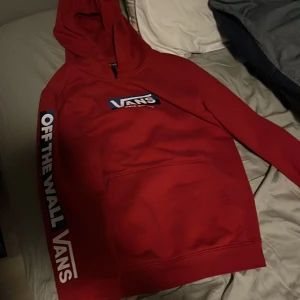 Röd hoodie från Vans - Säljer en snygg röd hoodie från Vans med tryck 'Off The Wall' på ärmen och logga på bröstet. Perfekt för en avslappnad stil. Hoodien har en klassisk känguruficka framtill.