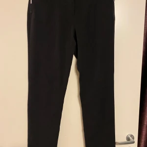 Brandtex Collection svarta byxor, storlek 40 - Eleganta svarta byxor från Brandtex Collection. Byxorna har detaljer som dragkedjor och dekorativa knappar fram och bak. Midjemått tvärsöver 42cm. Beninnerlängd 79cm.