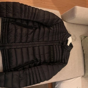 Svart pufferjacka från Moncler - Snygg svart pufferjacka från Moncler med dragkedja och långärmad design. Jackan har en quiltad stil och Moncler-logga på bröstet. Perfekt för kyligare dagar.