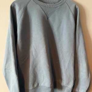 Grå sweatshirt från Carhartt - Säljer en stilren grå sweatshirt från Carhartt. Tröjan har en klassisk rund halsringning med ribbade muddar vid ärmslut och nederkant. Perfekt för en avslappnad look.