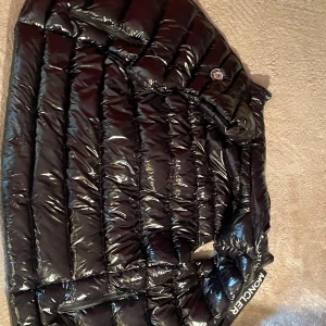 Svart glansig dunväst från Moncler - Säljer en svart glansig dunväst från Moncler med hög krage och dragkedja framtill. Västen har ett stilrent märke på bröstet och praktiska fickor med dragkedja. Perfekt för kyliga dagar när du vill hålla stilen.