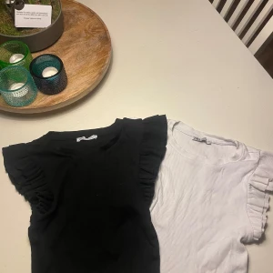 Svart och vit ribbad topp med volangärmar - Två snygga ribbade toppar i svart och vit med volangdetaljer på ärmarna. Perfekta för en stilren look. De är kortärmade och gjorda i ett mjukt material.