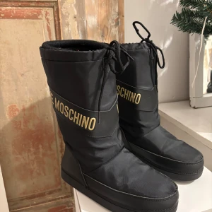 Svarta moon boots från Moschino - Snygga svarta moon boots från Moschino med guldiga detaljer och text. Skorna har en robust sula med bra grepp och snörning upptill. Perfekta för kalla vinterdagar med stil.Storlek 37/38
