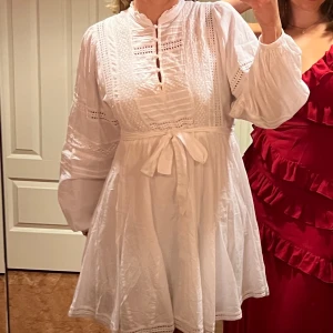 Vit klänning med knytband - Charmig vit klänning med knytband i midjan och vackra spetsdetaljer. Klänningen har långa ärmar och en kort längd, perfekt för en somrig look. Den har en rund halsringning med knappar framtill.