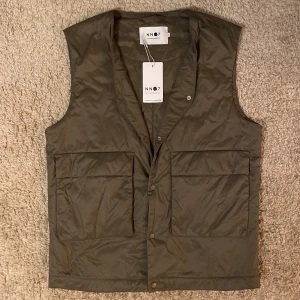 Olivgrön väst från NN07 - Snygg olivgrön väst från NN07 med praktiska fickor framtill. Västen har en stilren design med knappar och är perfekt för lager-på-lager-stil. Passar bra till både avslappnade och mer uppklädda tillfällen. Original tags medföljer.