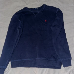 Mörkblå tröja från Polo Ralph Lauren - Säljer en snygg mörkblå tröja från Polo Ralph Lauren med det klassiska röda logotypbroderiet på bröstet. Tröjan har långa ärmar och en rund halsringning. Perfekt för en stilren och avslappnad look.
