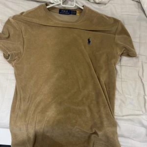 Beige t-shirt från Ralph Lauren - Säljer en stilren beige t-shirt från Ralph Lauren med det klassiska broderade logotypen i blått på bröstet. T-shirten är kortärmad och tillverkad i ett mjukt material som ger en lyxig känsla. Perfekt för en avslappnad och elegant look.