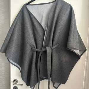 Grå kappa/ poncho med bälte och vid passform - Säljer en stilren grå kappa med bälte i midjan och vid, flowy passform. Jackan har öppen front och är perfekt för lager-på-lager. Enkel och snygg design som passar till många outfits. Perfekt för dig som gillar minimalistisk stil.
