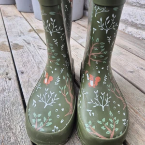 Gröna gummistövlar från Kuling strl 31 - Snygga gröna gummistövlar från Kuling med skogsmotiv och söta ekorrar. Stövlarna har mönster av träd, svampar och blad i vitt, brunt och grönt. Praktiska dragflikar baktill och grov sula för bra grepp. Perfekta för regniga dagar och äventyr utomhus. Hynd finns i hemmet.