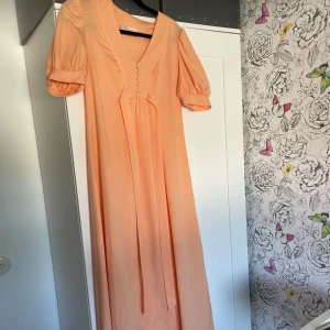 Persikofärgad långklänning med puffärm - Superfin långklänning i persikofärg med v-ringning, puffiga korta ärmar och klädda knappar framtill. Klänningen har ett knytband i midjan och är gjord i ett lätt, flowy material som ger lite retro vibe!