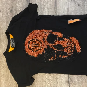 Svart t-shirt med orange döskalle Philipp Plein - Svart t-shirt från Philipp Plein med en stor orange döskalle framtill, dekorerad med små pärlor för en extra edgy look. Klassisk passform och rund halsringning. Perfekt för dig som vill sticka ut med en cool och unik stil.