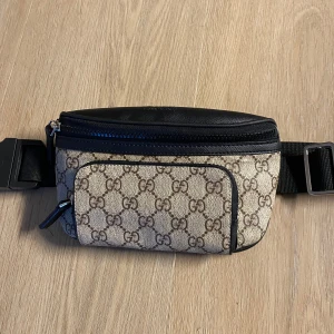 Gucci beige och svart magväska - Säljer en snygg magväska från Gucci med klassiskt GG-mönster i beige och brunt. Väskan har svarta skinn-detaljer, justerbart band och flera dragkedjefack. Perfekt för att bära det viktigaste med stil och attityd.