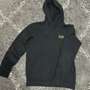 Svart EA7 hoodie med guldig logga - Svart hoodie från EA7 med guldig logga på bröstet och cool detalj i gult på ryggen. Tröjan har huva med snörning, känguruficka och långa ärmar. Perfekt för dig som gillar stilren streetwear med en touch av sport.