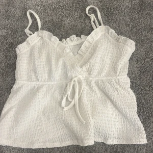 Vit linne från SHEIN - Supersöt vit linnetopp med omlott och knyt framtill. Toppen har smala axelband, volangkant längs urringningen och en lätt rynkad struktur i tyget. Perfekt för varma dagar och ger en romantisk vibe till din outfit.