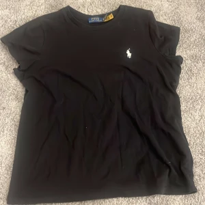 Svart t-shirt från Polo Ralph Lauren - Svart t-shirt från Polo Ralph Lauren med klassisk vit logga broderad på bröstet. Enkel och stilren design med rund halsringning och korta ärmar. Skön passform i mjuk bomull, perfekt till jeans eller shorts.