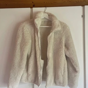 Beige teddyjacka med dragkedja - Supermjuk och fluffig teddyjacka i beige med hög krage och dragkedja framtill. Perfekt för kyliga dagar när du vill vara både varm och snygg. Jackan har lång ärm och en avslappnad passform som ger en chill vibe.