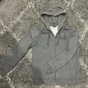 Grå hoodiejacka från Emporio Armani - Grå hoodiejacka från Emporio Armani EA7 med dragkedja, huva med snörning och stora logotryck på ryggen. Jackan har två fickor fram och är tillverkad i mjuk bomullsmix. Perfekt för en chill och sportig look.