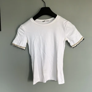 Vit t-shirt med spetsdetalj från Zara - Vit t-shirt från Zara i bomull med korta ärmar. Ärmarna har snygga spetsdetaljer i svart, vitt och guld som ger en unik touch. Perfekt för dig som gillar stilrena plagg med det lilla extra. Normal passform och rund halsringning.