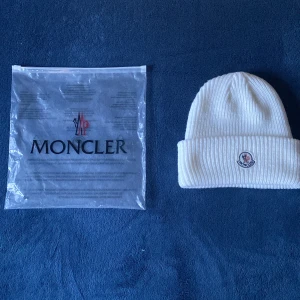 Vit ribbstickad mössa från Moncler - Vit ribbstickad mössa från Moncler med klassisk uppvikt kant och broderad logotyp framtill. Mössan är tillverkad i mjukt material och har en clean, stilren look som passar perfekt till kalla dagar. Snygg detalj med Moncler-tag baktill.