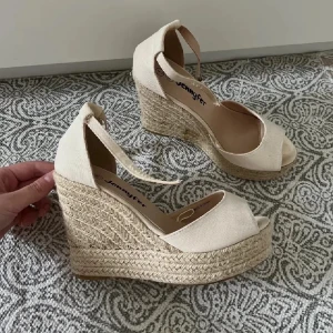 Beiga espadrille wedges från Jennyfer - Snygga beiga espadrille wedges från Jennyfer med öppen tå och knytband runt ankeln. Skorna har hög flätad plattformsula i jute och ovandel i textil. Perfekta för en somrig look med extra höjd och stil.