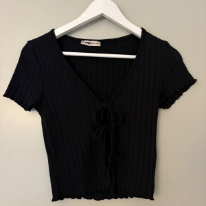 Svart ribbad knyt-topp från newyorker - Svart croppad topp med ribbad struktur och v-ringning. Korta ärmar och snygg knytning framtill ger en trendig look. Perfekt till jeans eller kjol för en cool stil.