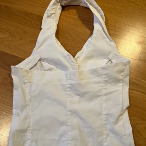 Vit halterneck topp med knappar - Snygg vit topp i halterneck-modell med krage och fyra knappar framtill. Den har två fuskfickor och markerade sömmar som ger en figurnära passform. Perfekt för att lyfta en outfit med en clean och fräsch look. Hör av dig vid frågor, lägg prisförslag ❤️