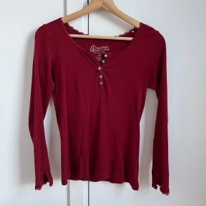 Vinröd långärmad topp med spetsdetaljer - Supersöt vinröd långärmad topp från Esprit i mjuk ekologisk bomull. V-ringad med knappar framtill och fina spetsdetaljer vid halsringning och ärmslut. Perfekt för dig som gillar en feminin och chill stil. 💓