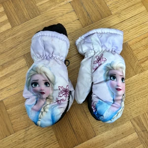 Frozen-vantar med lila snöflingor - Säljer ett par söta Frozen-vantar med motiv av Elsa och lila snöflingor. Vantarna är tumhandskar med svart grepp på handflatan och mjukt svart fleecefoder inuti. Perfekta för kalla vinterdagar och för alla som älskar Frost!