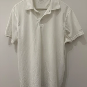 Vit piké t-shirt från AONE - Klassisk vit piké t-shirt från AONE i storlek L. Tröjan har korta ärmar, krage och knäppning framtill. Tillverkad i snabbtorkande material som passar perfekt för aktiva dagar. Enkel och stilren design med diskret logga på ärmen.