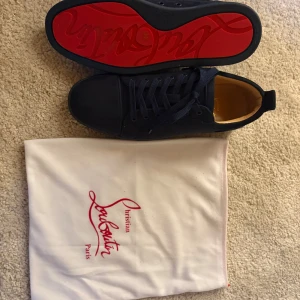 Christian Louboutin svarta mockasneakers - Snygga svarta sneakers från Christian Louboutin i mocka med klassisk röd sula och diskret logga. Skorna har rund tå, platt sula och snörning. Perfekta för dig som vill ha en lyxig och stilren look med en ikonisk touch.