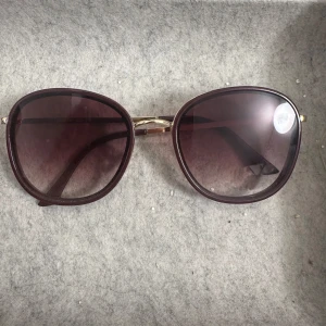 Bruna solglasögon från Lindex - Stilrena solglasögon från Lindex med stora, runda bågar i mörkbrunt och guldiga detaljer vid skalmarna. Glasen är tonade i en mörk nyans som ger en cool vibe. Perfekta för att lyfta din look under soliga dagar.