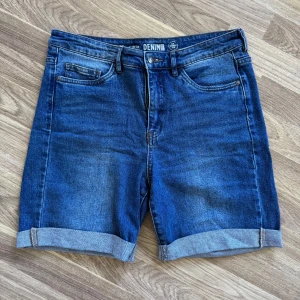 Blå jeansshorts från Denim 1953 - Snygga blå jeansshorts från Denim 1953 med klassisk femficksdesign och uppvikta benslut. Modellen är midwaist/highwaist och har bälteshällor samt knapp- och dragkedjestängning. 